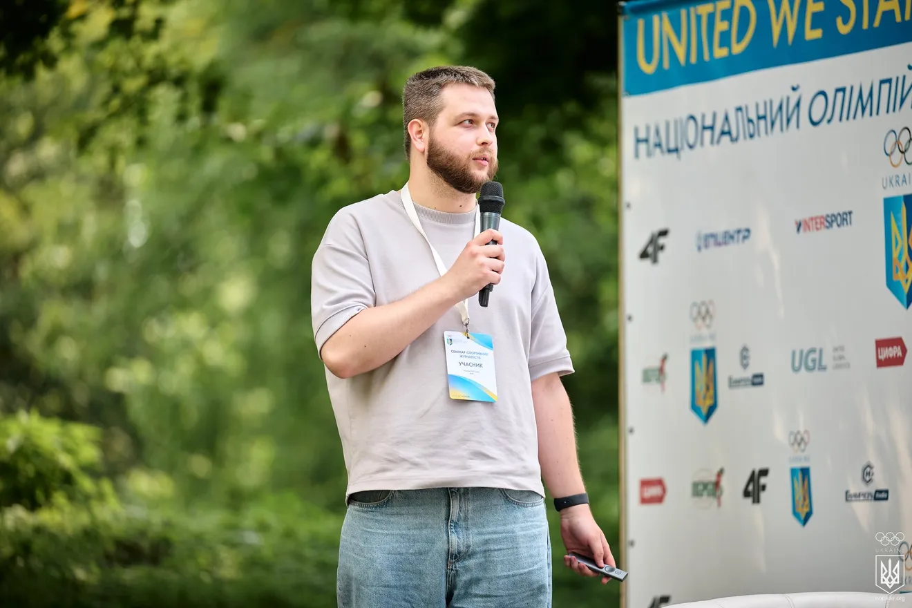Богдан Щербаков говорить у мікрофон просто неба, поруч банер Національного олімпійського комітету України з олімпійськими кільцями