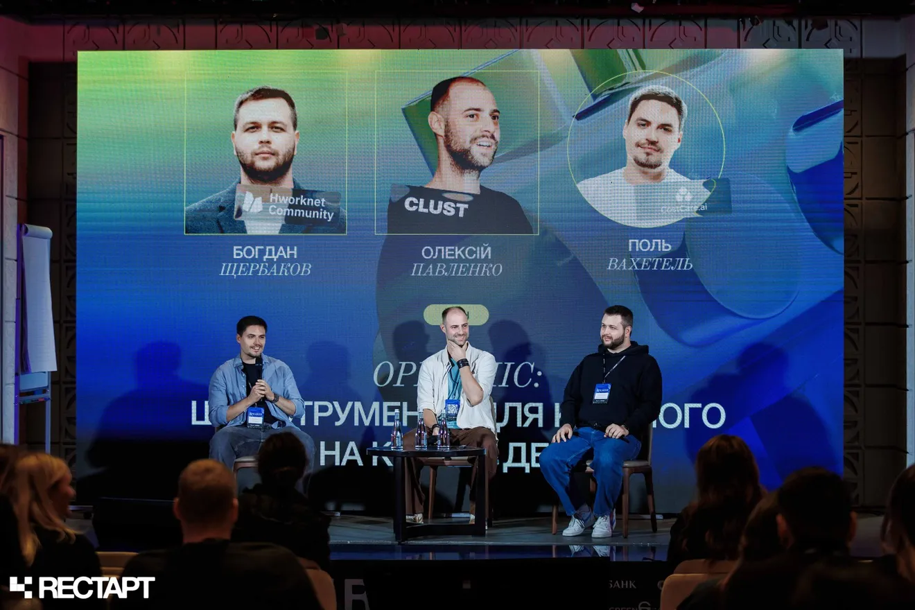 Богдан Щербаков праворуч на сцені під час панельної дискусії з двома спікерами, на екрані позаду — їхні портрети, у першому ряду глядачі